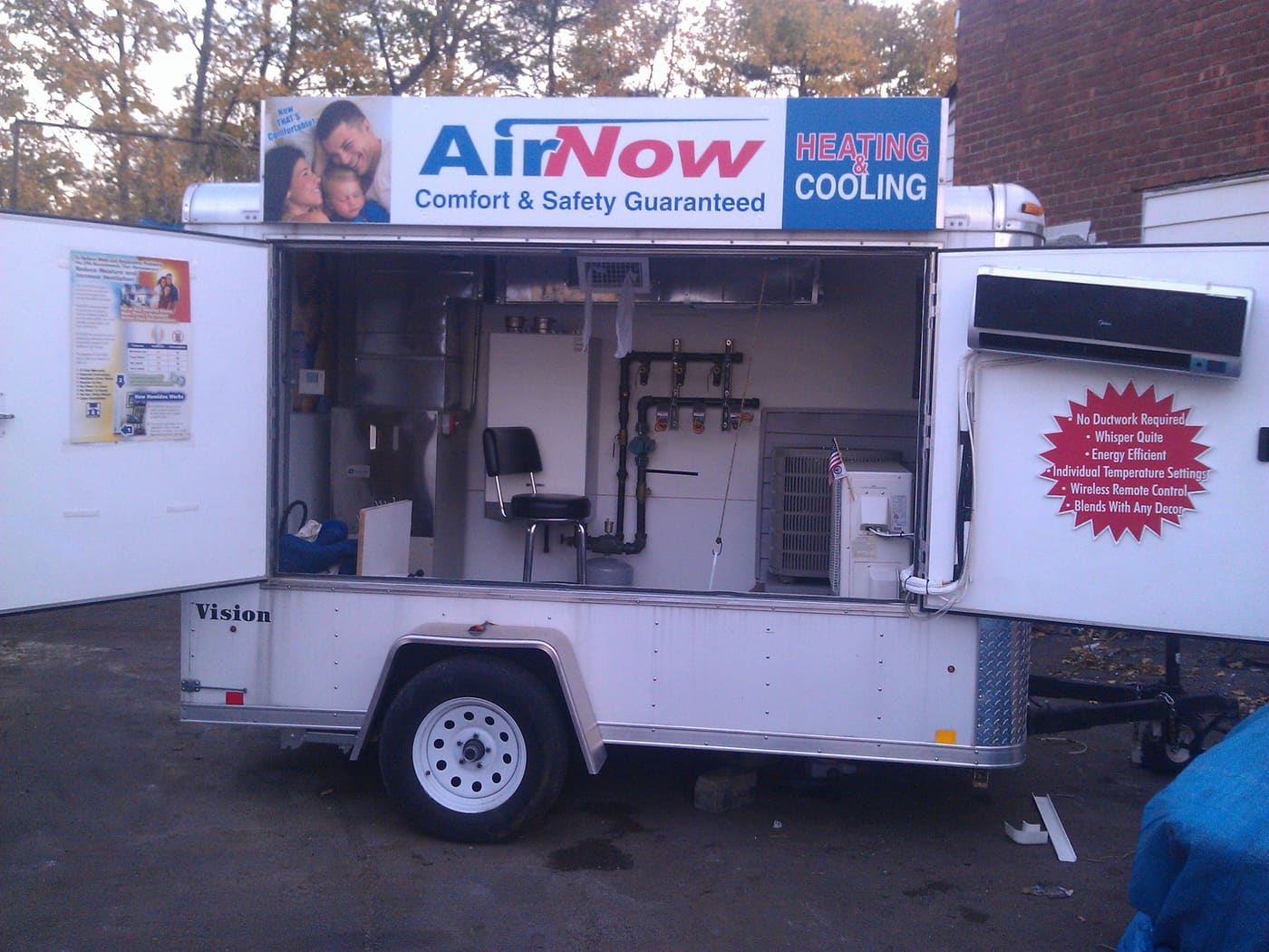 A white single-axle event trailer with its side panels swung open, displaying HVAC equipment inside &mdash; an air handler, copper piping and manifolds, a condenser unit and a wall-mounted ductless head &mdash; under signage reading AirNow Heating & Cooling.